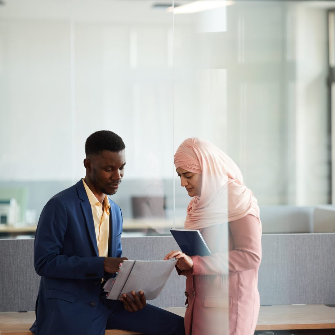 Vertical portrait of elegant African-American man talking to young Muslim businesswoman and holding documents in modern office interior, copy space