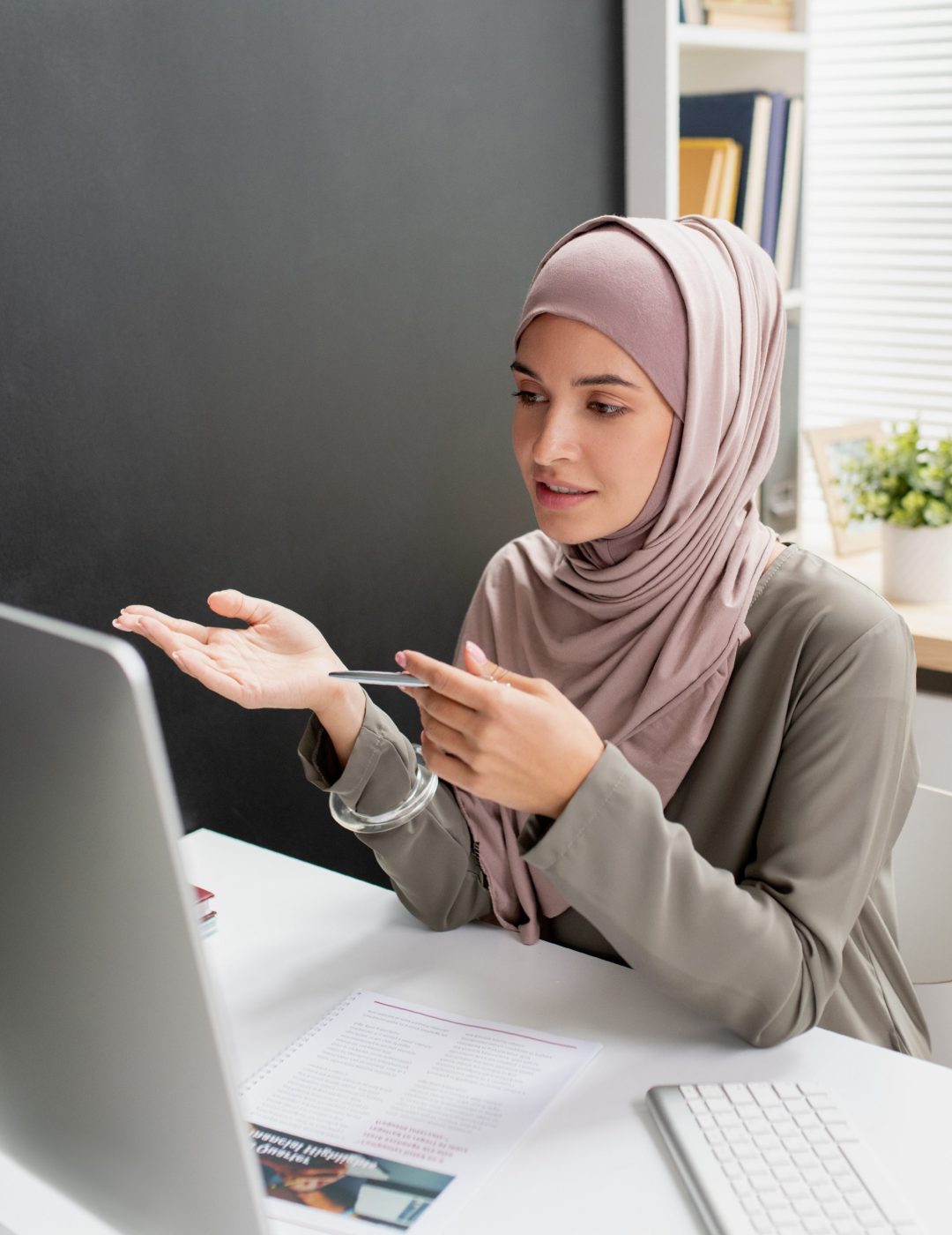 Young confident teacher in hijab speaking to online audience in front of computer monitor while sitting by desk in home environment